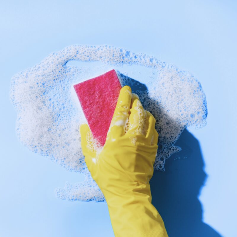 Gelber Handschuh reinigt mit Schwamm eine schaumige Fläche auf blauem Hintergrund.