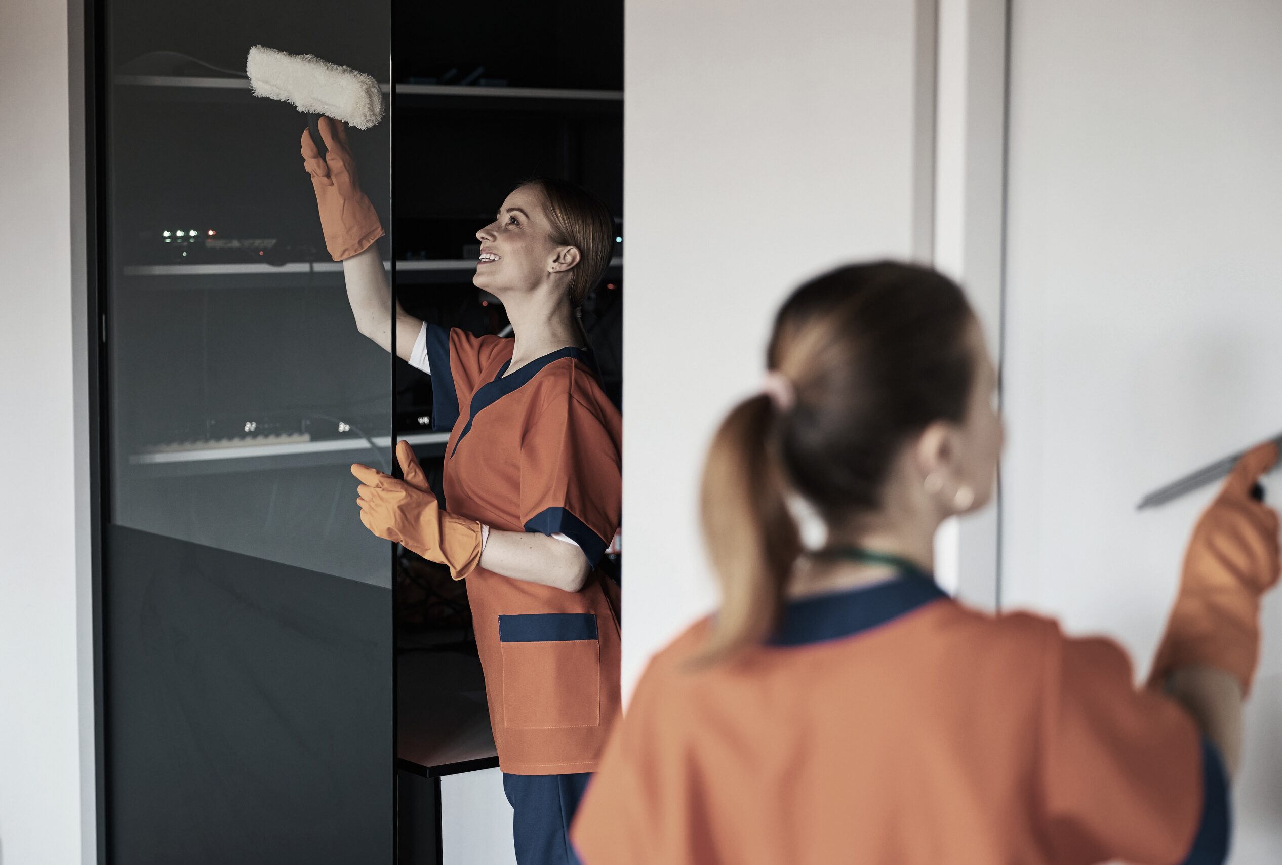 Reinigungskraft reinigt Glasflaeche mit Einwascher, zweite Person im Vordergrund.